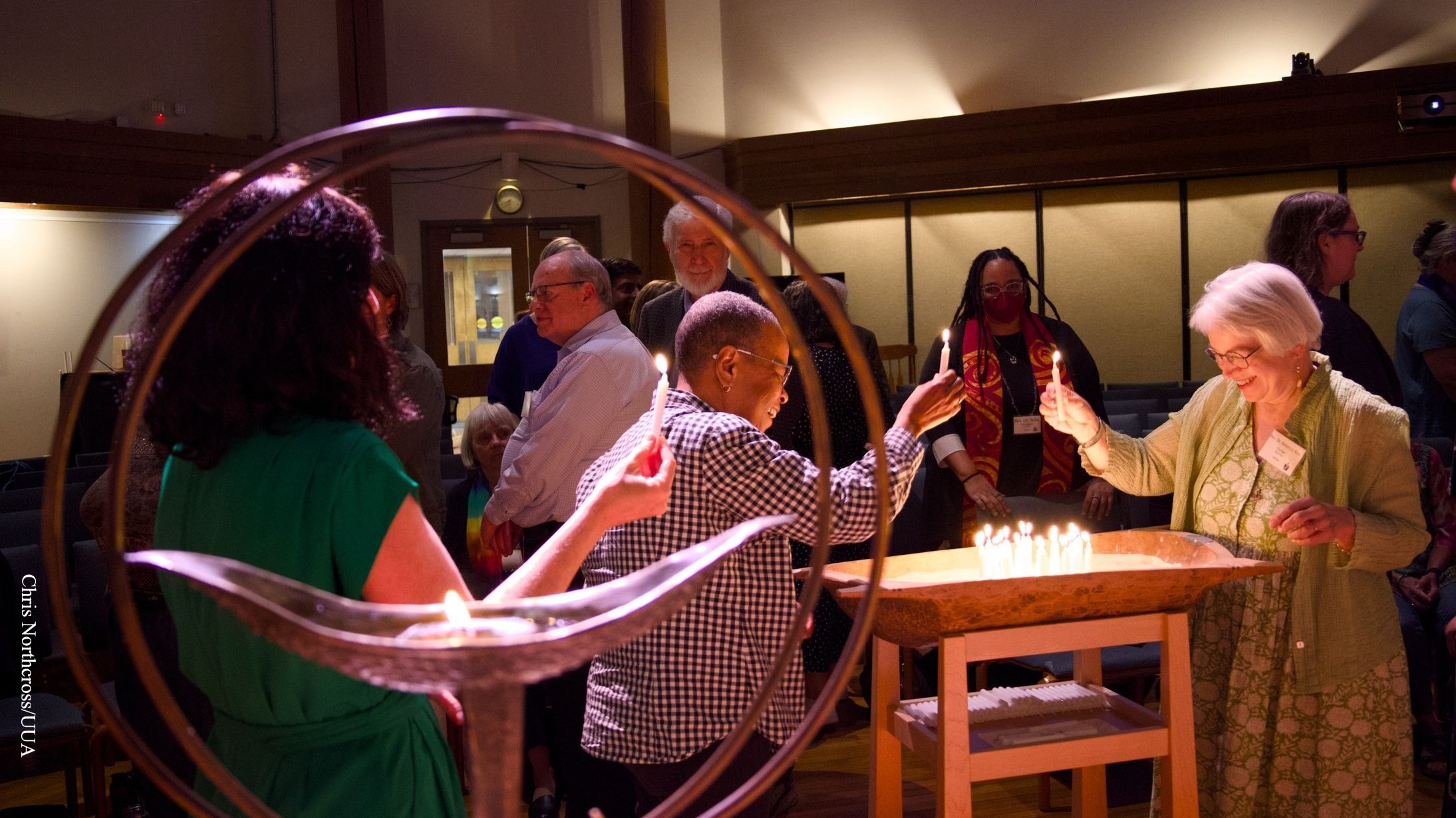 An image of diverse people gathered in a large, low-lit sanctuary, talking and lighting candles that they will then set on a table with other lit candles. A brass, 2-ringed UUA chalice is near the camera and half the image is seen through the rings.