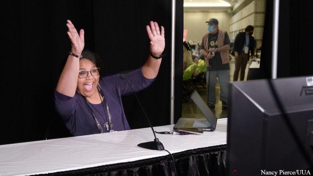 A person of color wearing glasses and a dark shirt is at a table in the GA delegate booth, smiling broadly and waving both hands. A microphone is set in front of them and a laptop and computer monitor can be seen from an angle at the table. In the background, a person wearing a baseball cap, striped vest, and a mask can be seen through a doorway, walking among others who are seated and standing.
