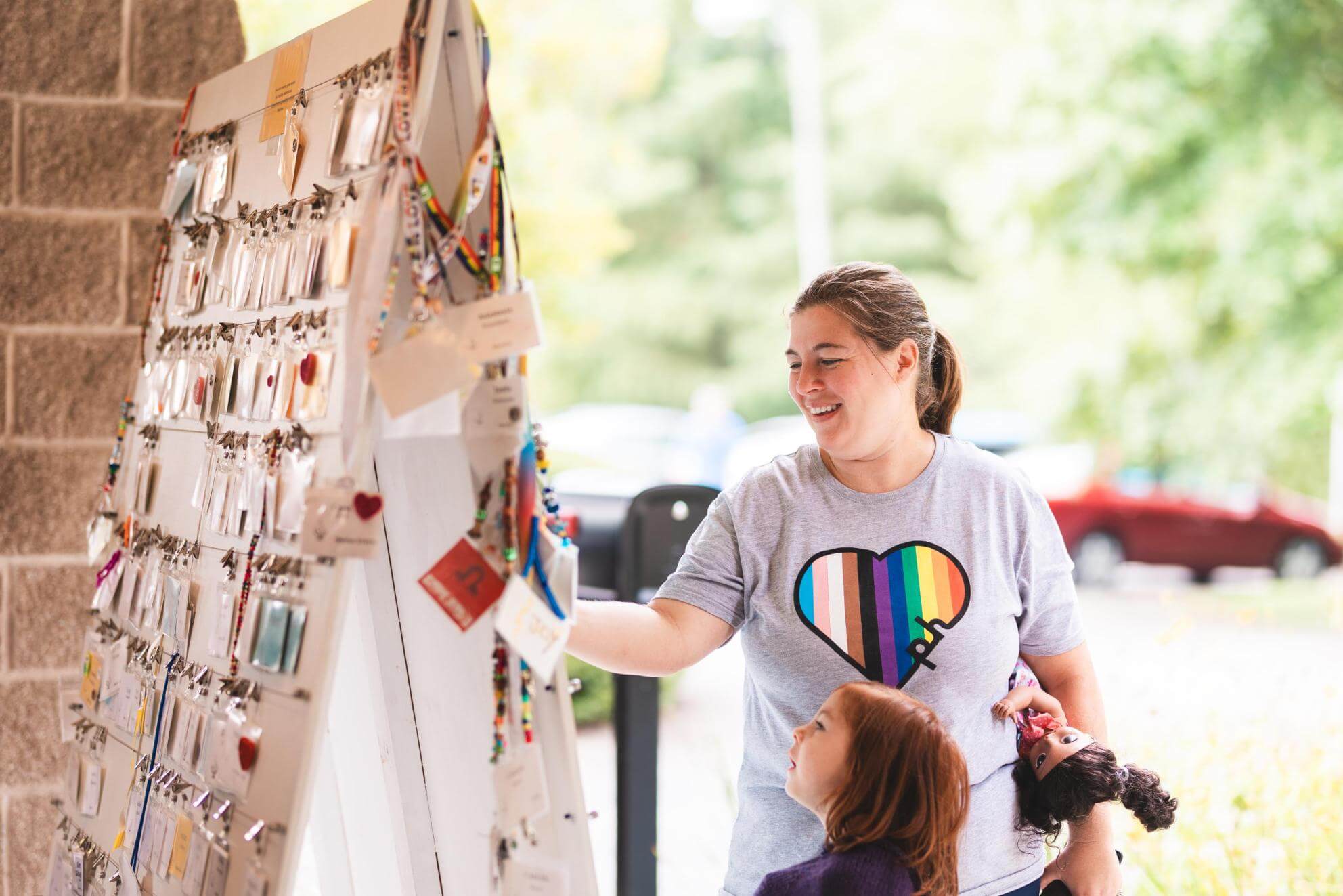 A person wearing a shirt with a rainbow pride flag-striped heart on it stands next to a child and takes a nametag off of a rack at a Unitarian Universalist congregation. 